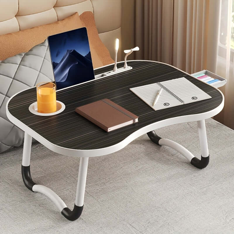 Portable laptop bed table with modern design, featuring a cup holder, notebook, and laptop on a bed.