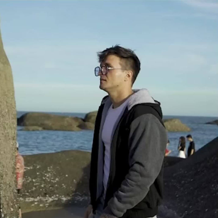 A man wearing a men's hooded jacket specifications stands by the beach looking into the distance.
