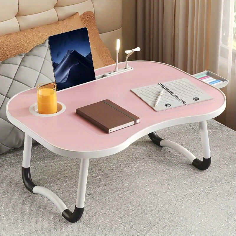 Portable laptop bed table with pink surface, laptop, notebook, and cup, perfect for comfortable work from bed.