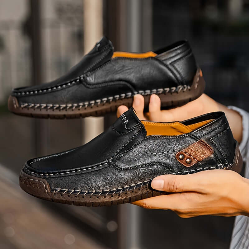 men's slip-on loafers in black leather being held by a person, showcasing detailed stitching and comfortable design
