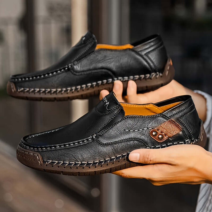 men's slip-on loafers in black leather being held by a person, showcasing detailed stitching and comfortable design