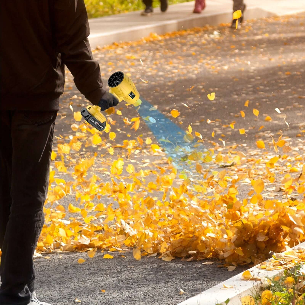 Person using a leaf blower to clear yellow autumn leaves from a sidewalk in a vibrant fall setting.