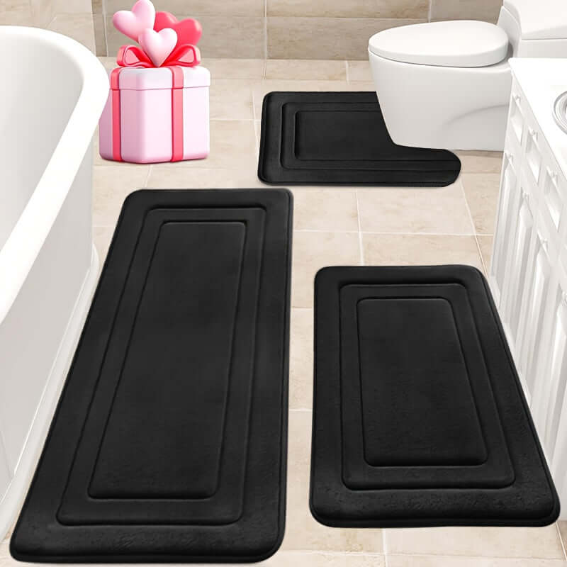 Memory foam bath mat set in black on a tile bathroom floor with a gift box nearby.