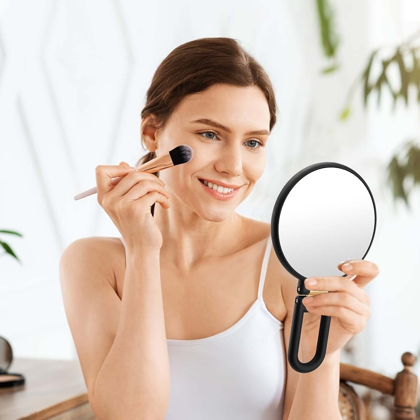 Woman applying makeup with a brush while using a 50x magnifying handheld mirror.